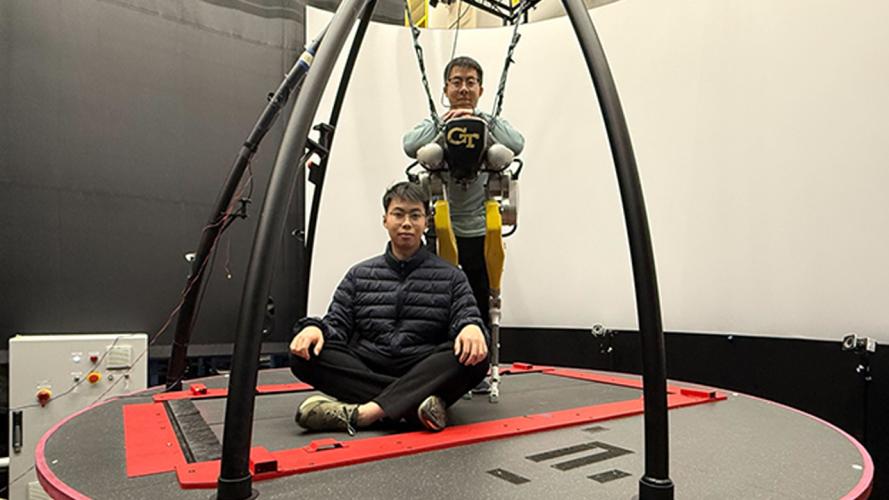 Two individuals on a large circular motion platform in a research lab, with one person seated cross-legged on the platform and another suspended in a harness wearing a Georgia Tech exoskeleton system.