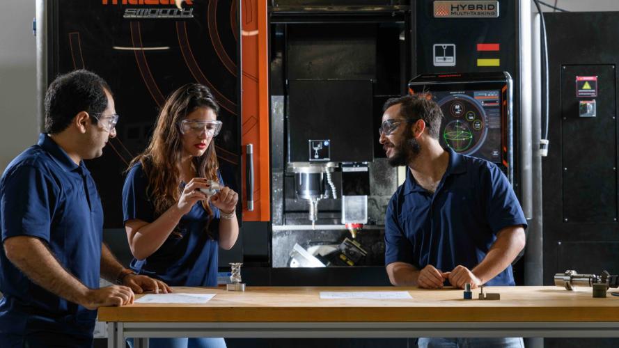 Three individuals standing at a workbench in front of a large Mazak hybrid manufacturing machine, discussing components and technical drawings.