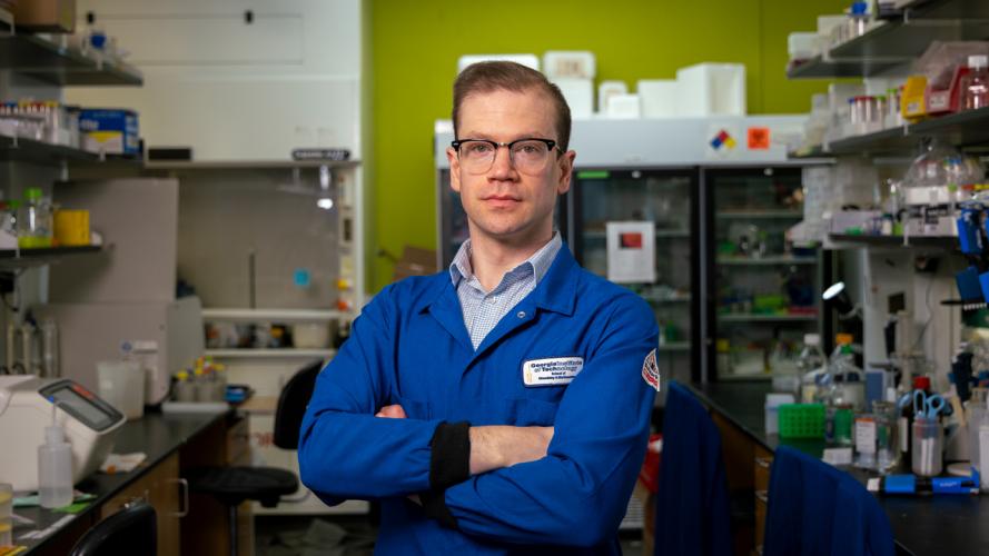 Researcher in a blue lab coat stands with arms crossed in a laboratory surrounded by scientific equipment and storage units.