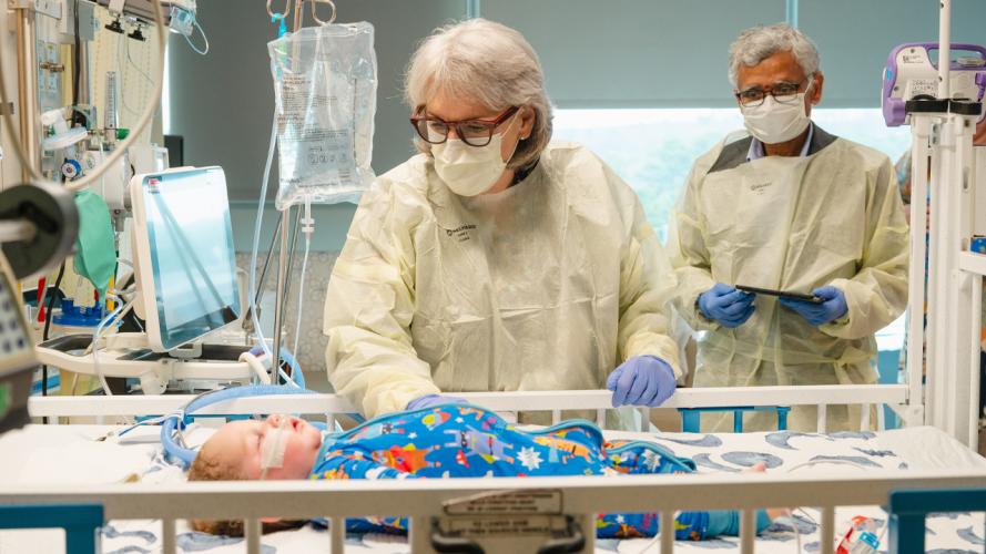 Medical researchers in protective gowns and masks observe a child patient in a hospital bed surrounded by monitoring and medical equipment.