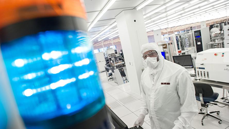 A cleanroom technician in protective gear works at a computer workstation in a semiconductor lab, with a blue signal light in the foreground and lab equipment behind them.