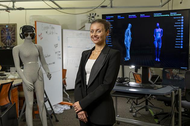 female with her hair pulled up and wearing a black suit jacket standing in front of two screen monitors showing blue outlines of human forms