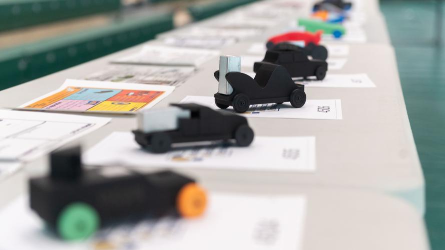 A row of small, student‑designed model race cars displayed on a table, each placed on top of design sheets in a gym setting.