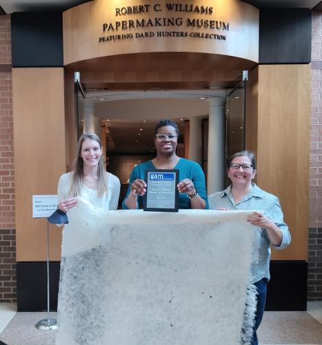 Three museum staff stand in front of the museum doors holding a very large sheet of paper and a plaque. 