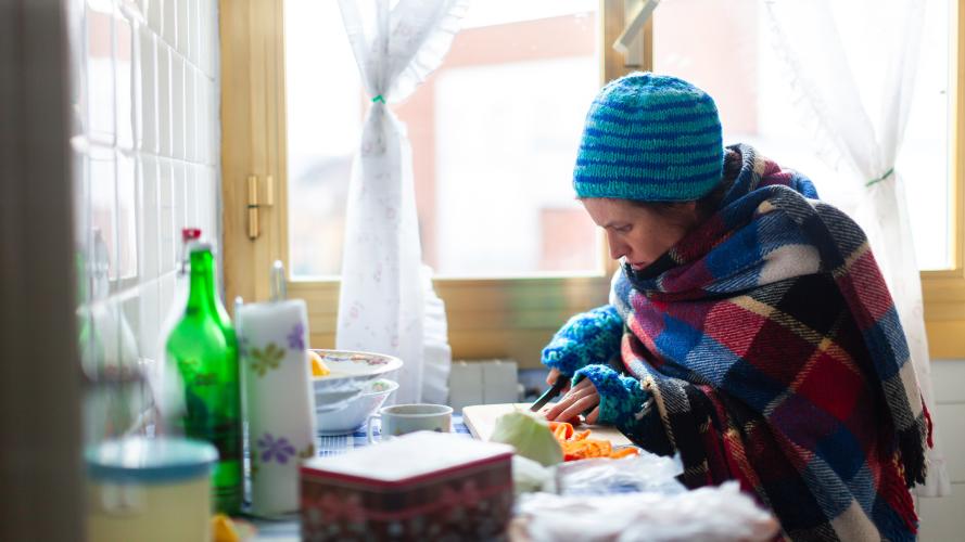 A woman wearing a hat and warm clothing prepares food in her kitchen.