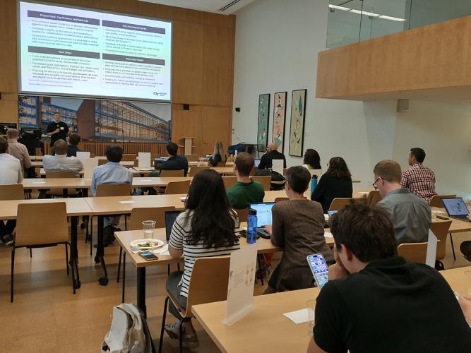 View of a man presenting to a crowd of onlookers in a large meeting room.