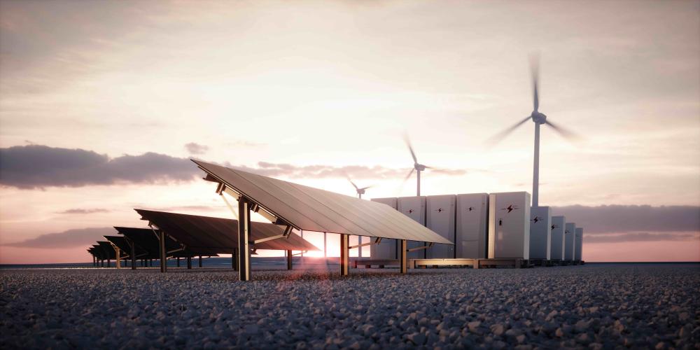 Ground‑mounted solar panel array in the foreground with wind turbines and large battery storage units visible in the background under a cloudy sky.