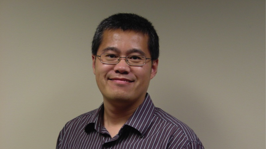 Portrait of a smiling man with short dark hair and glasses wearing a striped button-down shirt against a plain beige background.