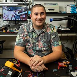 Josiah Hester sits at a desk in an electronics lab at Georgia Tech with an array of prototype projects and test equipment in front of him.