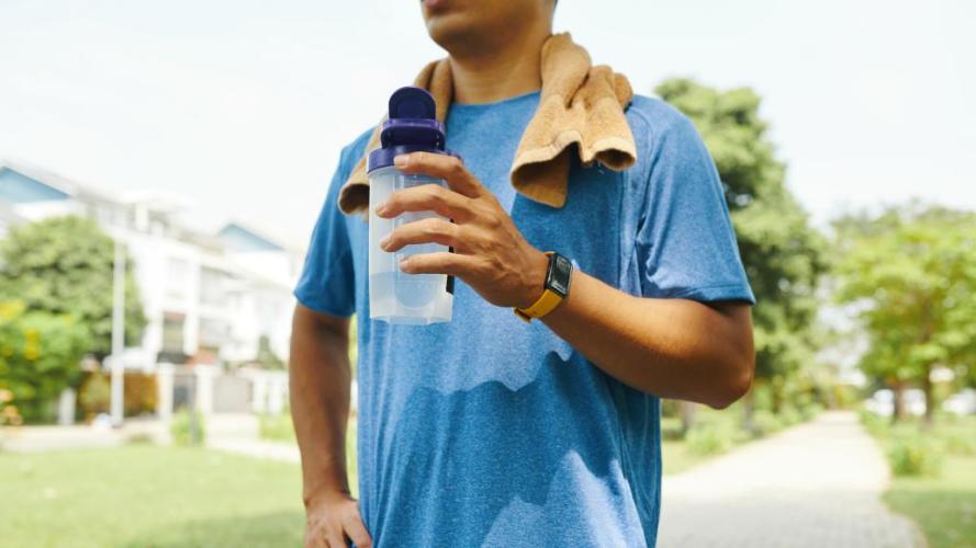 Person holding a water bottle in their left hand wearing a smart watch and a towel on his shoulder appearing to be after a workout