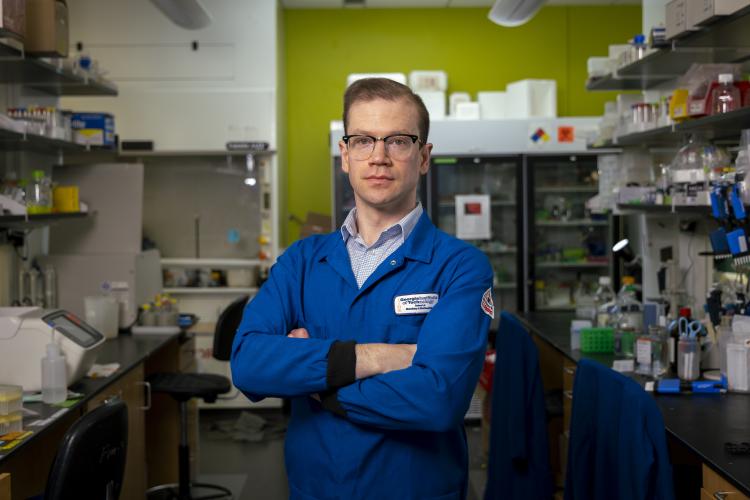 A person wearing a blue lab coat stands with arms crossed in a laboratory filled with shelves of scientific equipment, supplies, and a refrigerator unit in the background.