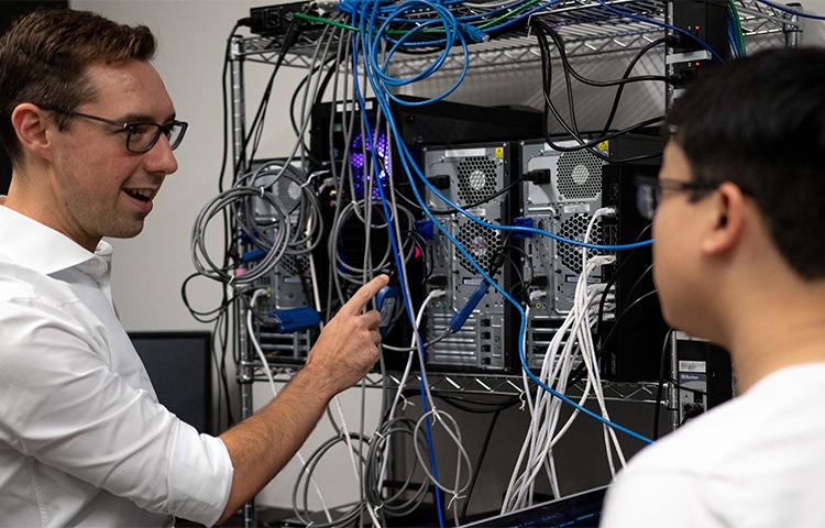 A man points to a rack of CPUs. He is talking to a student who is slighlty out of focus. 