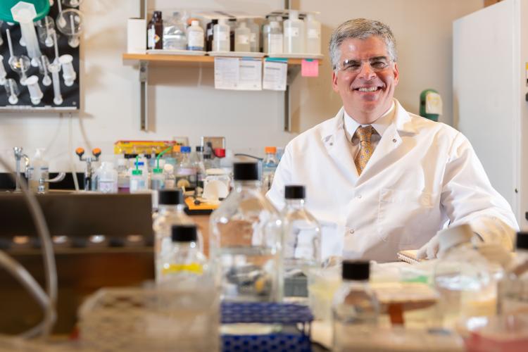 A man with silver hair wears a white lab coat, white shirt, and gold tie will sitting behind a lab bench with research equipment on top of it.