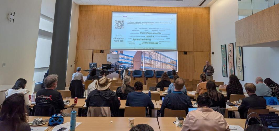 A view inside the Scholars Event Theater of a session of the Sustainability Showcase. A man speaks to a crowd while presenting slides on a large projection screen.