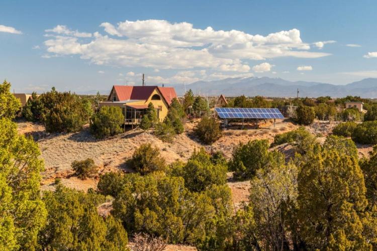 A rural residence with solar panels installed outdoors, set among desert vegetation with mountains in the distance.
