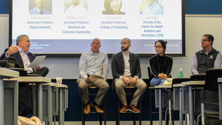 A panel of five speakers sits on tall stools at the front of a classroom, participating in a moderated discussion. The moderator on the left holds papers while addressing the group. A large presentation slide behind the panel displays names and academic titles. Audience members are partially visible in the foreground, and tables, chairs, and a water bottle are arranged throughout the room.