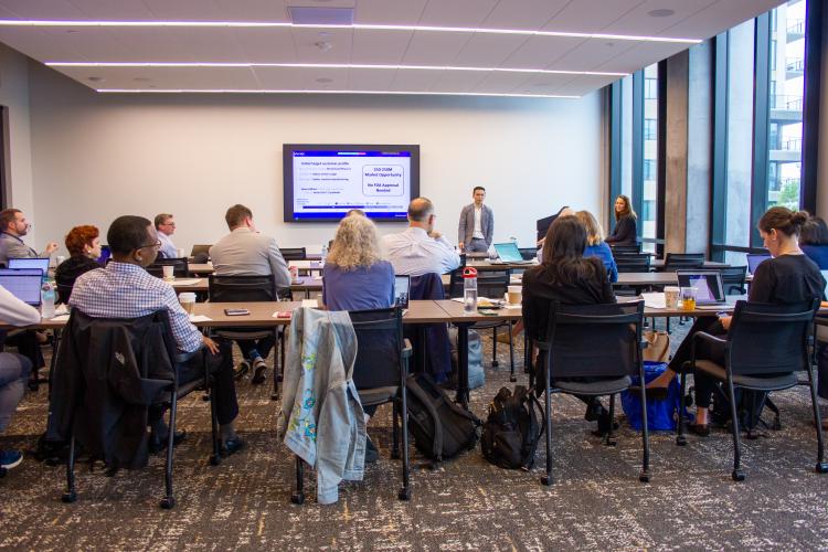 A photo shot from the back of a conference room with people sitting at conference tables while a person at the front of the room shows a presentation on a flat TV screen