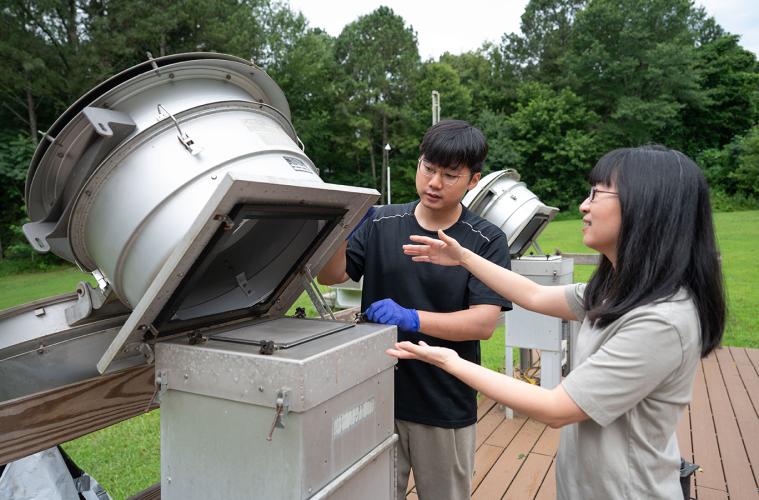 A male and female researcher working with a metal piece of equipment outdoors with trees and grass in the background