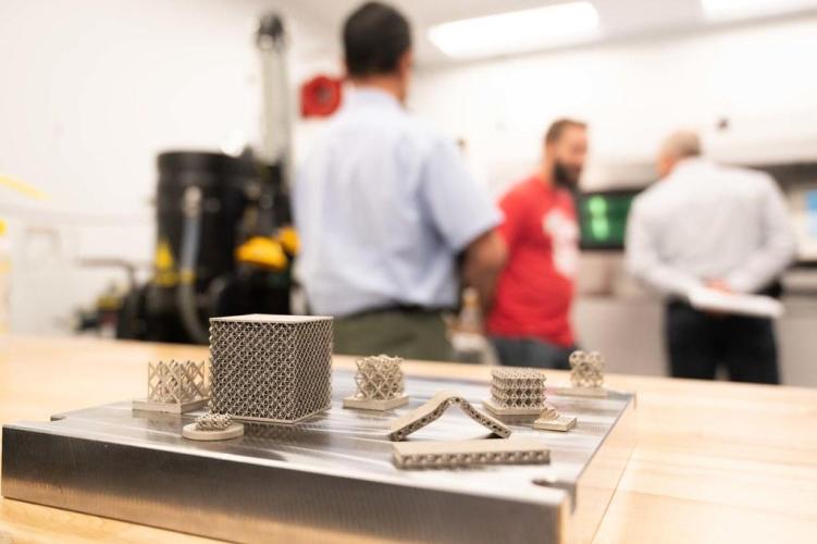 Small metal lattice and cylindrical components arranged on a flat surface in the foreground, with several people standing and talking in a laboratory or workshop space in the background.
