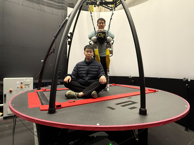 Two individuals on a large circular motion platform in a research lab, with one person seated cross‑legged on the platform and another suspended in a harness wearing a Georgia Tech exoskeleton system.