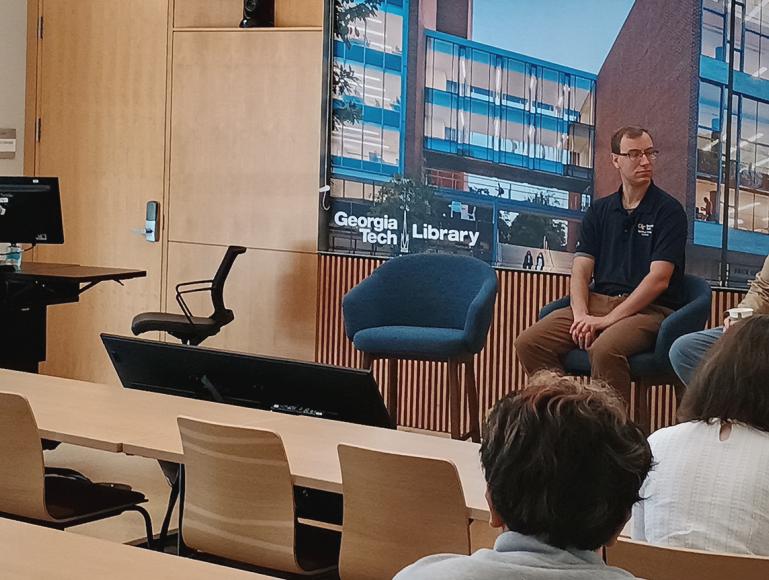 Panel discussion in a lecture room featuring two seated speakers at the front of the room and a presenter standing beside a lectern. A large display behind the speakers shows an image of the Georgia Tech Library building, while audience members are seated at tables facing the discussion. The room has light wood paneling throughout the space.