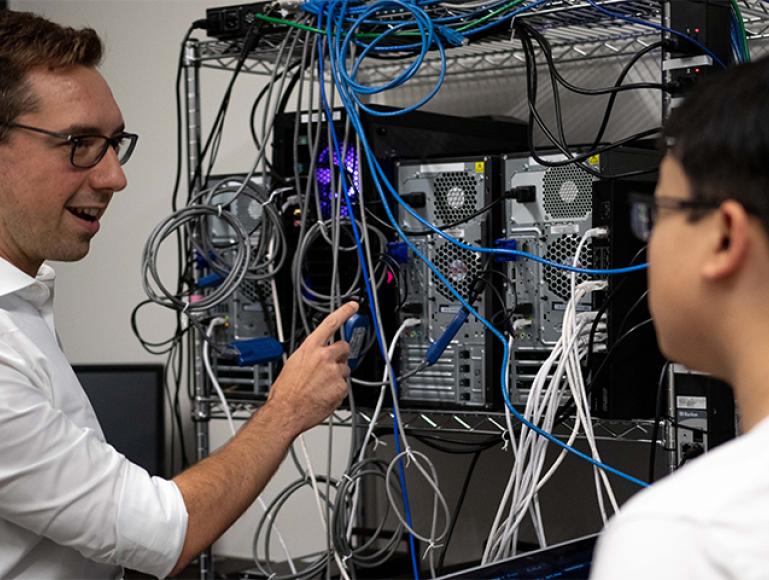 A man points to a rack of CPUs. He is talking to a student who is slighlty out of focus. 