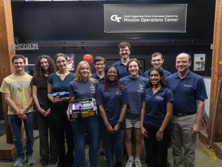 Georgia Tech students, faculty, and staff pose with spacecraft hardware beneath the Daniel Guggenheim School of Aerospace Engineering Mission Operations Center sign.