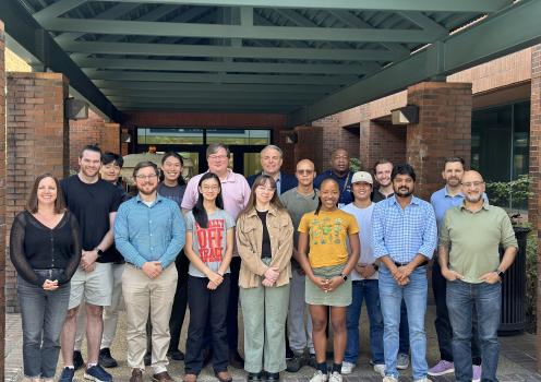 A group of students, researchers, and staff stand together for a group photo outside a brick building beneath a covered walkway.