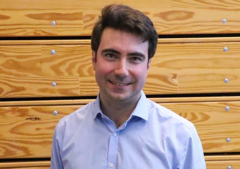 A man standing in front of a wooden wall. He is smiling and tilting his head. 
