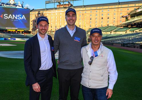 Three men who co-founded and lead Huxley Medical stand together on the field of a baseball stadium, smiling at the camera. They are wearing SANSA-branded hats, with a large video board behind them displaying the SANSA logo and the stadium seating in the background.