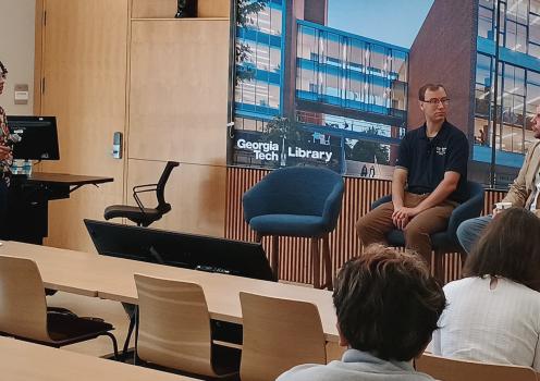 Panel discussion in a lecture room featuring two seated speakers at the front of the room and a presenter standing beside a lectern. A large display behind the speakers shows an image of the Georgia Tech Library building, while audience members are seated at tables facing the discussion. The room has light wood paneling throughout the space.
