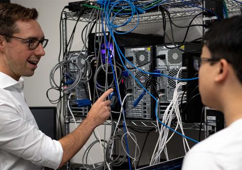 A man points to a rack of CPUs. He is talking to a student who is slighlty out of focus. 