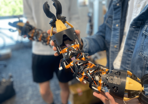 Close-up of a modular, snake-like robotic device with exposed wiring, sensors, and claw-like grippers at both ends being held by two people in a laboratory setting. The flexible robotic structure features black and orange components connected by segmented joints.