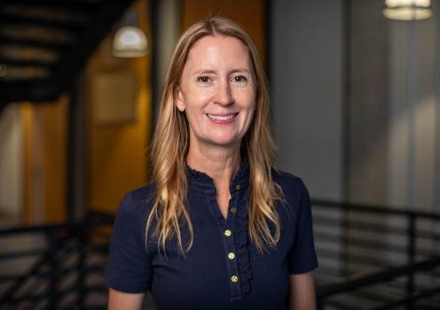A blond woman wearing a navy blue blouse smiles for a portrait with a staircase in the background