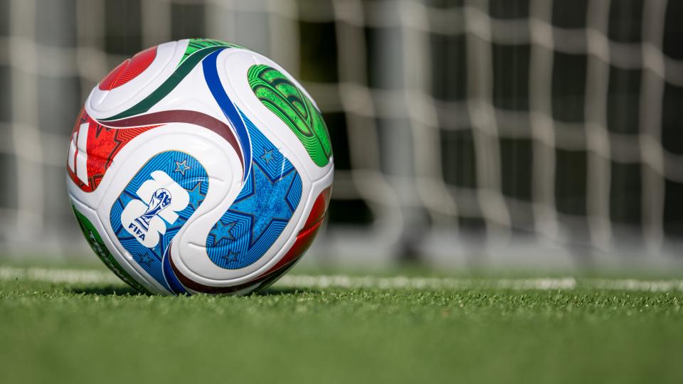 Close-up of a colorful FIFA soccer ball resting on a grass field with a blurred goal net in the background. The ball features blue, green, red, and white geometric designs and the FIFA logo.