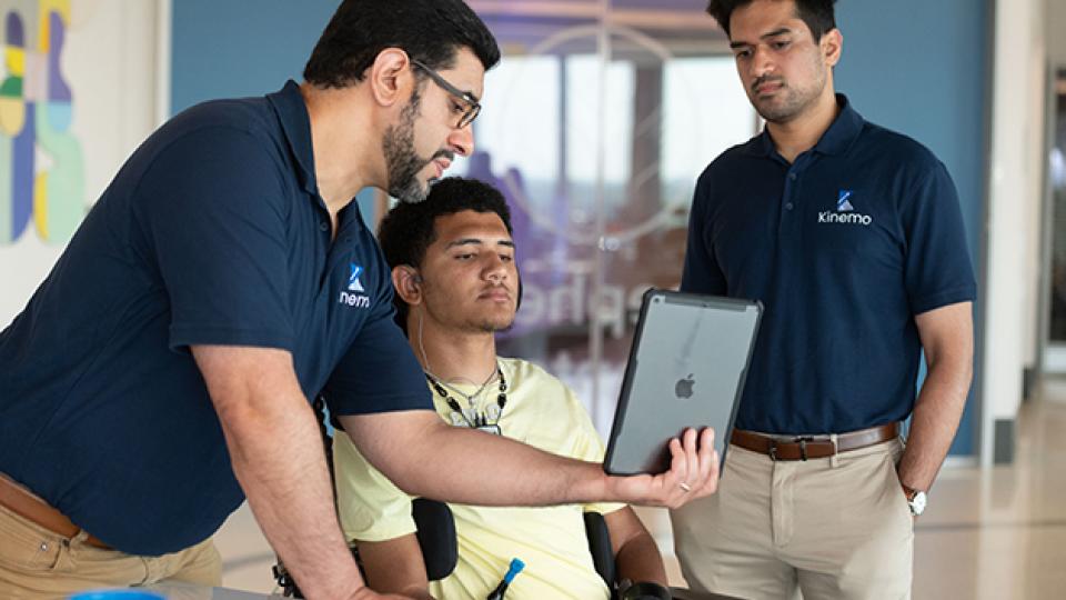 Three people review a tablet together in a lab setting, with one seated individual using a powered wheelchair and assistive device.