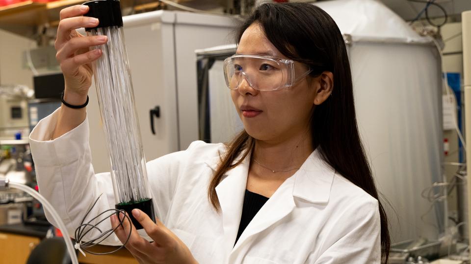 A woman with dark hair holds a glass tube filled with white fibers. 