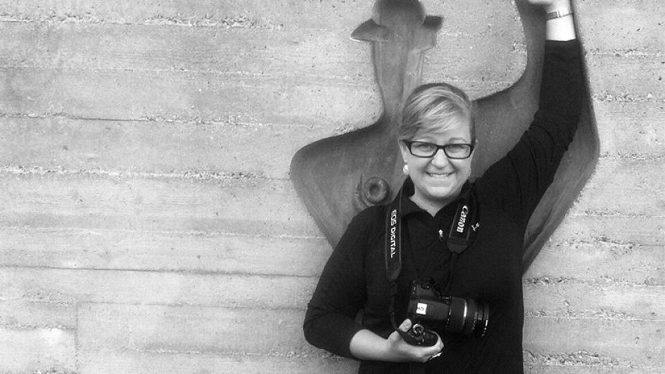 A woman holding a camera stands in front of a relief carving in a concrete wall mimicking its posture, with her arm raised over her head.