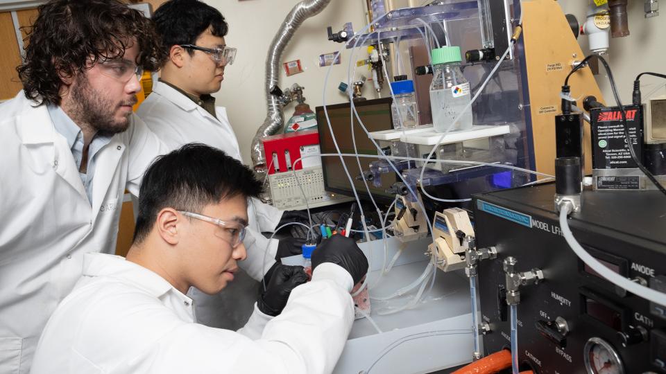 Three men in lab coats working at a bench on an experimental setup with tubes, vials, and pumps.