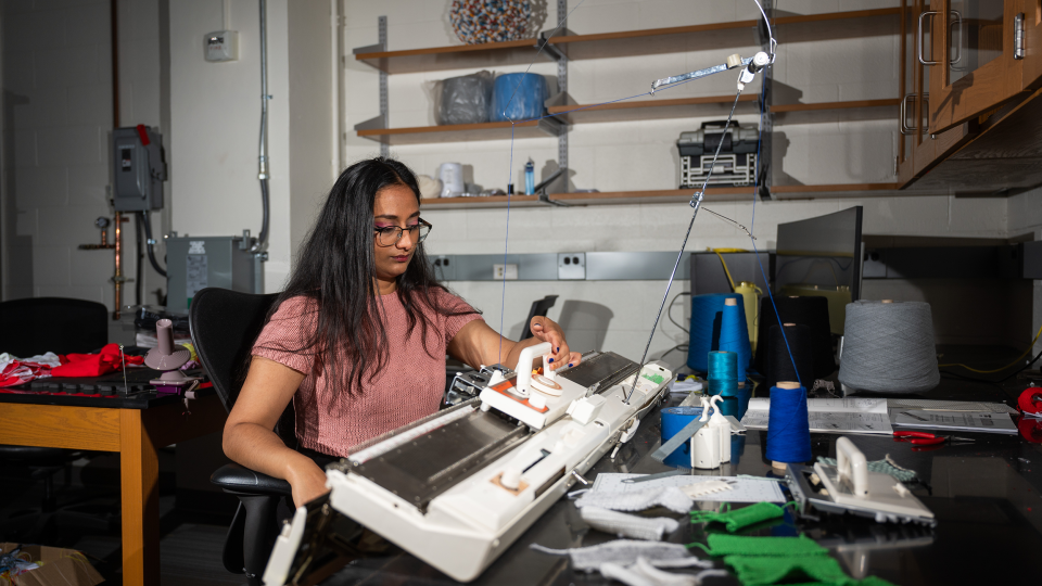 A woman wearing glasses and short sleeve pink sweater sit nexts to a commercial knitting machine.