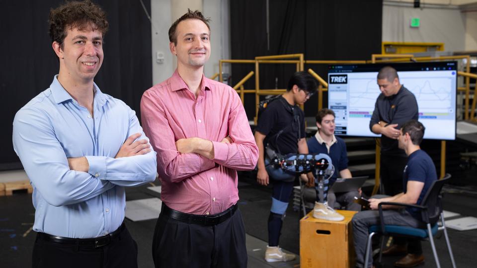 Matthew Gombolay and Aaron Young pose in the lab while Ph.D. researchers work on a leg exoskeleton device.