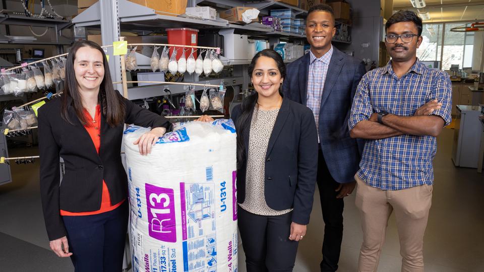 Four researchers standing in a lab with a large roll of fiberglass insulation and a wooden rack holding small bags of hemp fiber-based insulation materials. (Photo: Candler Hobbs)