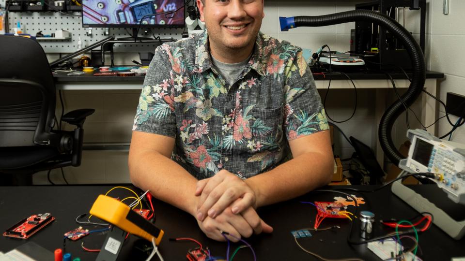Josiah Hester sits at a desk in an electronics lab at Georgia Tech with an array of prototype projects and test equipment in front of him.