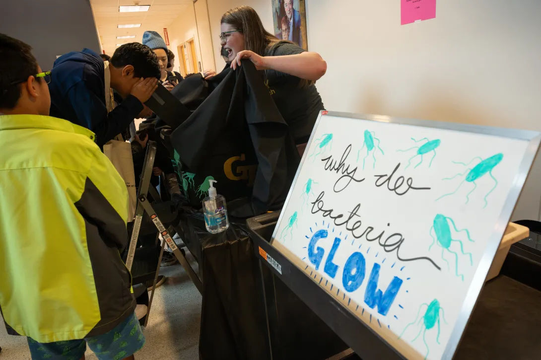 A person looks into a microscope. A sign with the text "why do bacteria GLOW" is in the foreground