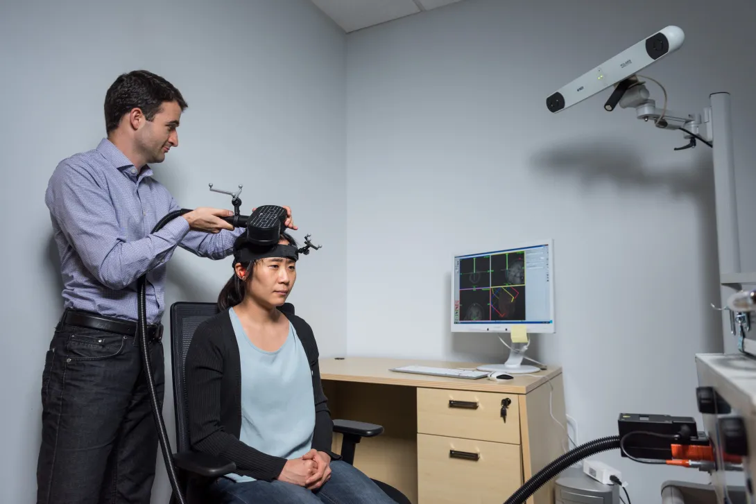 Researcher adjusting a device on another person’s head in a lab, with a computer displaying brain imaging data and a mounted camera in the background.