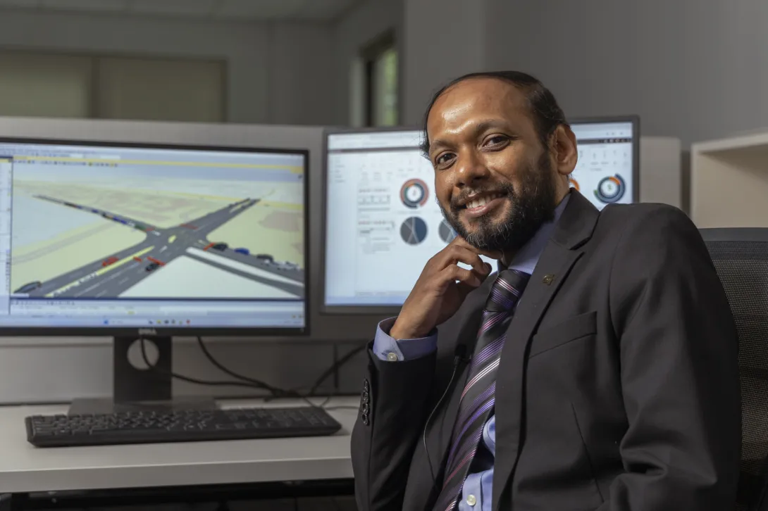 Angshuman Guin (a male professor wearing a black suit) sits at a desk in front of two monitors displaying data
