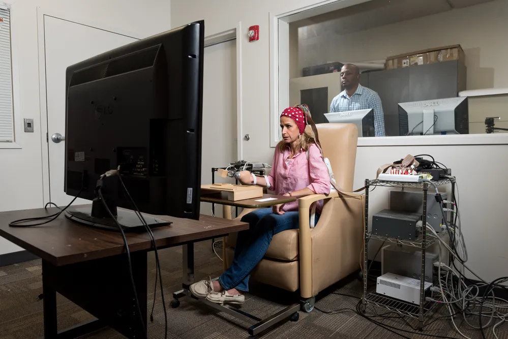 A person seated in a beige chair using a computer setup with multiple cables and devices, facing a large monitor in a testing or research room, with another individual visible through a window in an adjacent control room.