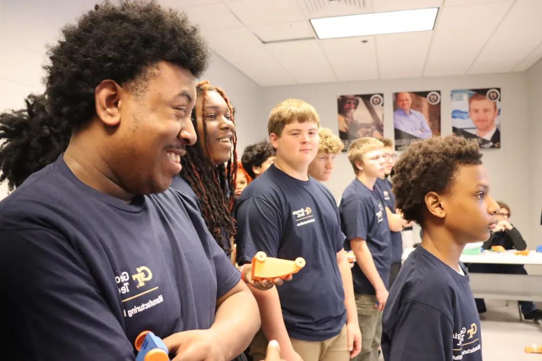 Group of students in navy shirts standing in a classroom, each holding small orange and blue 3D-printed objects.