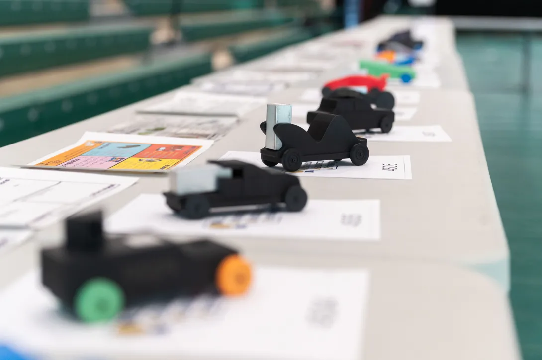 A row of small, student‑designed model race cars displayed on a table, each placed on top of design sheets in a gym setting.