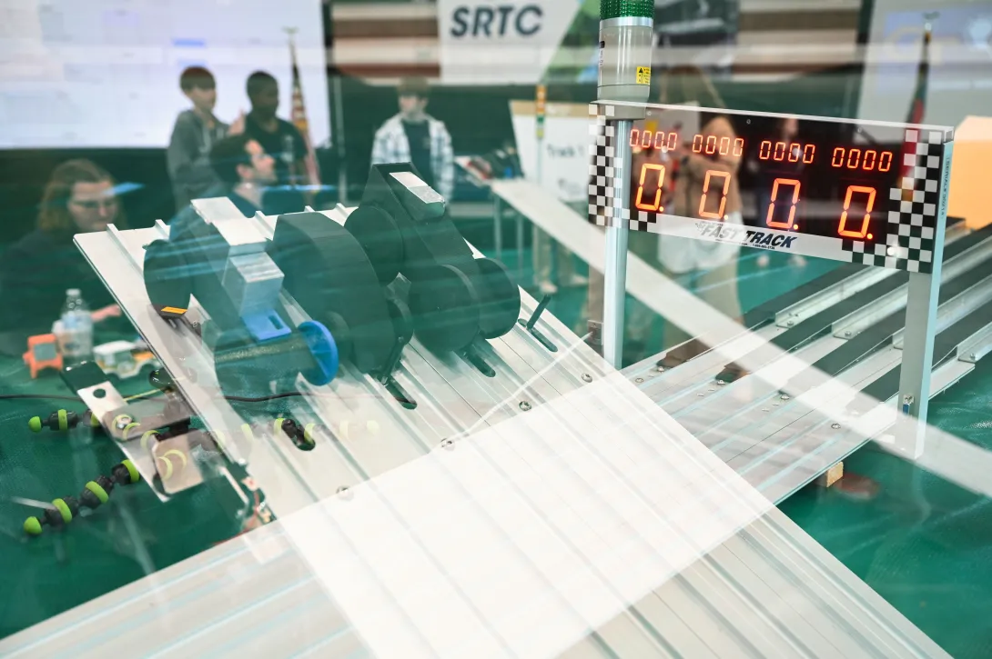 wo small student‑designed race cars positioned at the starting line of a metal track, with a digital timer display and spectators in the background at an Advanced Manufacturing competition.